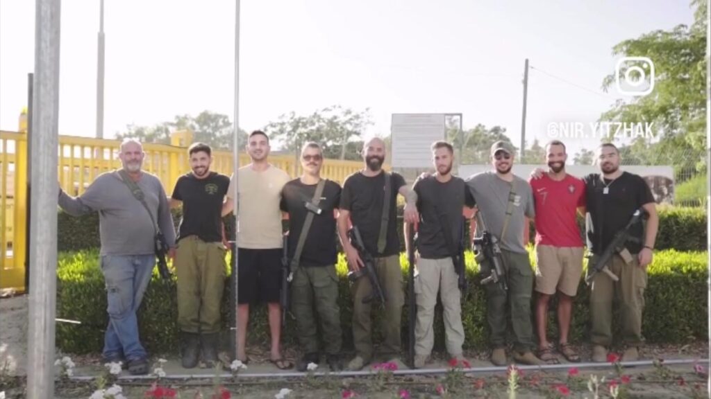 A line up of nine men, some of whom have rifles slung over their shoulders, in front of bushes and a yellow gate. 